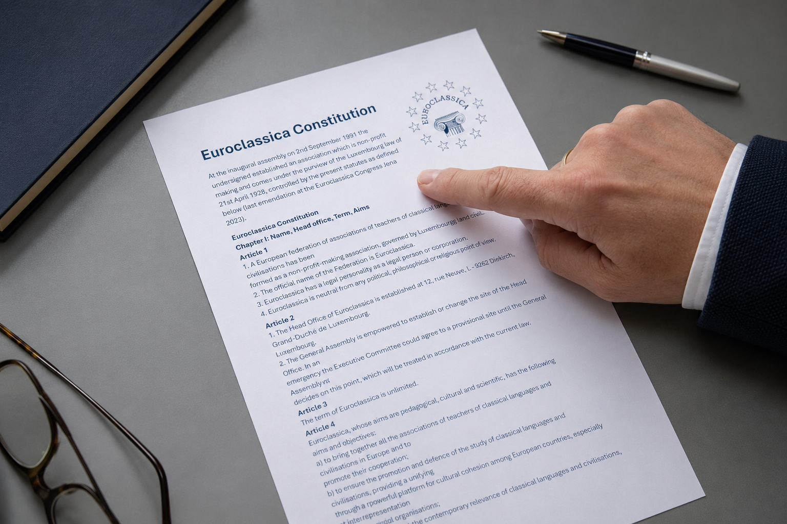 Hand pointing to the Euroclassica Constitution document on a desk with glasses, a notebook, and a pen.