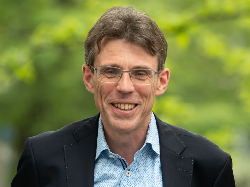 Portrait of Christian Laes smiling outdoors, wearing glasses, a dark blazer, and a light blue shirt with a blurred green background.