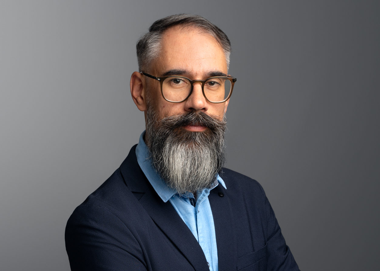Portrait of Šime Demo wearing a navy blazer and light blue shirt, standing with crossed arms against a grey background.